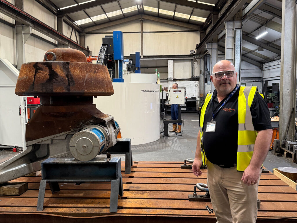 Leader CNC Technologies team member in factory standing next to a large component with two Shibaura Machine vertical borers behind